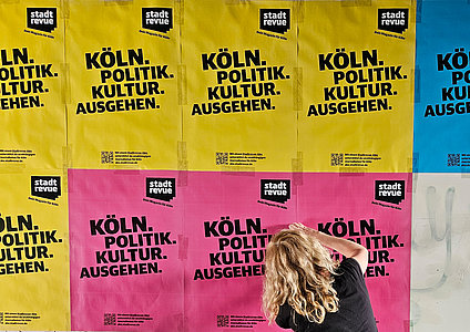 Eine Frau beklebt eine Wand mit bunten Plakaten. Darauf steht: "Köln, Politik, Kultur, Ausgehen — Stadtrevue, dein Magazin für Köln"