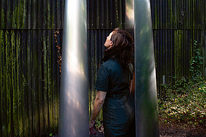 Künstlerin in der eigenen Installation stehend: Olga Holzschuh, songs of conditions, 2024, Schwarze Erde, Aluminium, Kupfer.